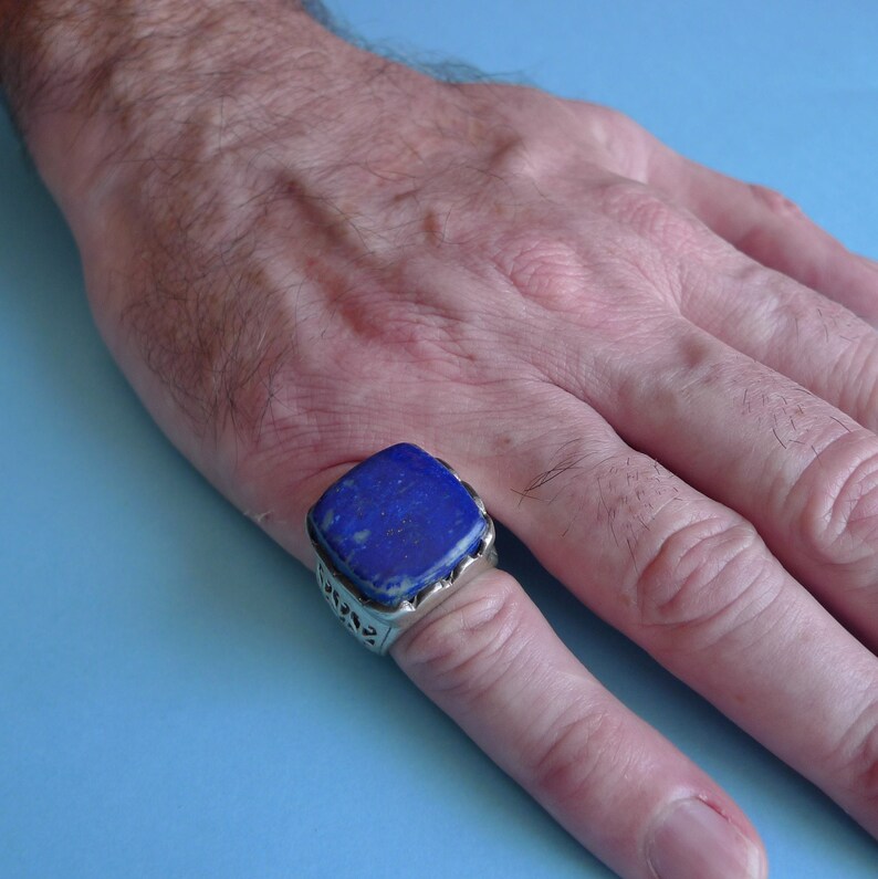 May include: A silver ring with a large, square, blue lapis lazuli stone set in the center. The ring is worn on a person's finger.