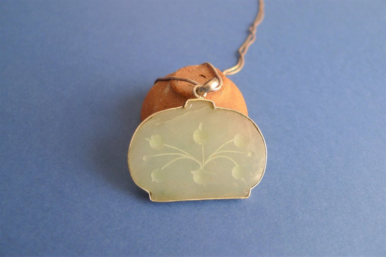 May include: A green jade pendant necklace with a floral design. The pendant is oval shaped and has a silver bail.