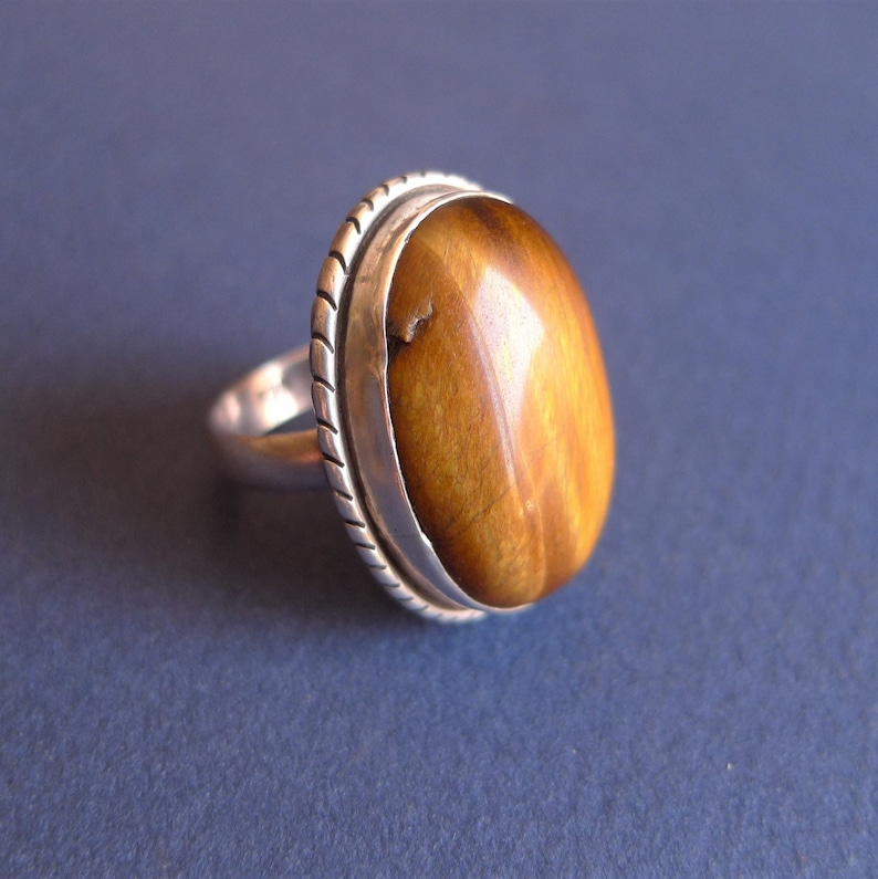 K&ouml;nnte beinhalten: Ein silberner Ring mit einem braunen und goldenen Tigerauge-Stein, der in einem seilartigen Band gefasst ist.