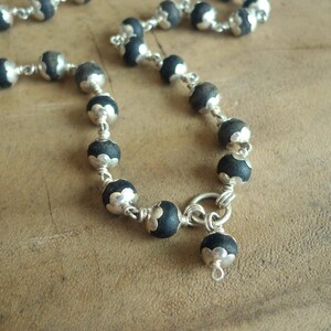 May include: A close-up of a necklace with black beads and silver-coloured accents. The beads are round and linked by a silver chain. The necklace is on a brown surface.