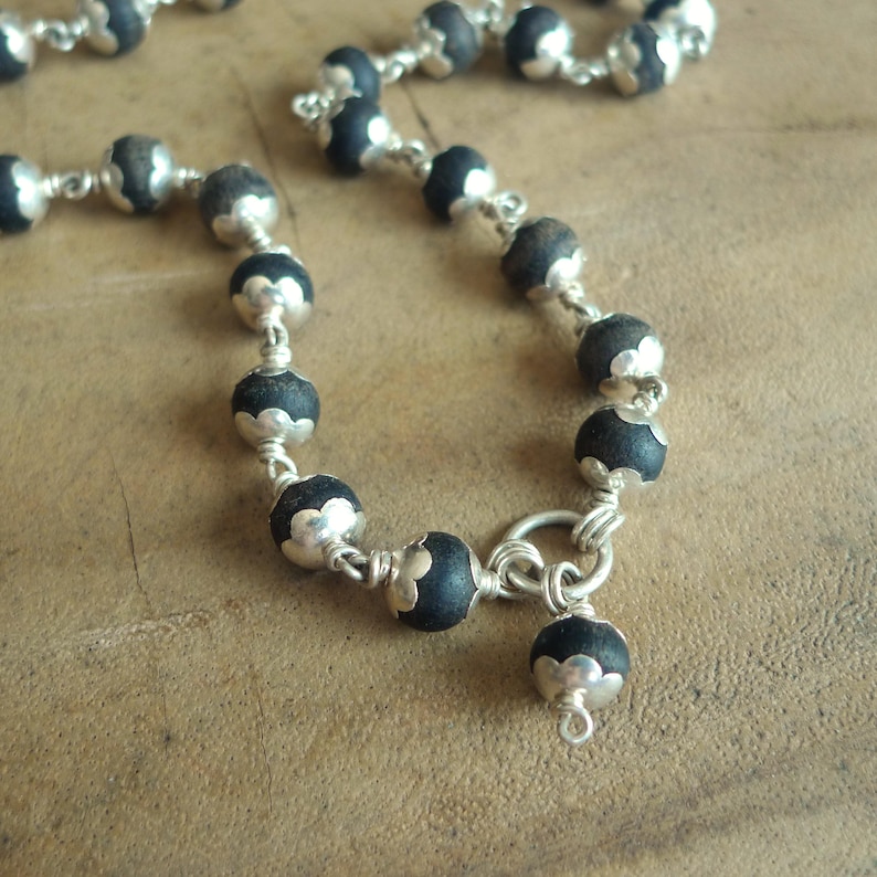 May include: A close-up of a necklace with black beads and silver-coloured accents. The beads are round and linked by a silver chain. The necklace is on a brown surface.