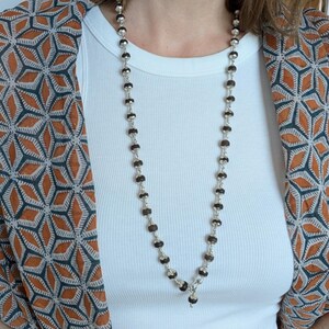May include: A long necklace with dark brown, faceted beads and silver-coloured metal beads. The necklace is worn over a white ribbed vest top. A patterned orange, brown, and grey kimono-style jacket is worn over the vest top.