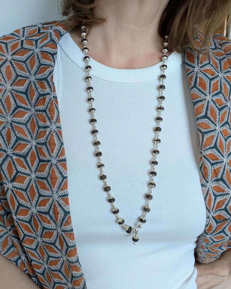 May include: A long necklace with dark brown, faceted beads and silver-coloured metal beads. The necklace is worn over a white ribbed vest top. A patterned orange, brown, and grey kimono-style jacket is worn over the vest top.