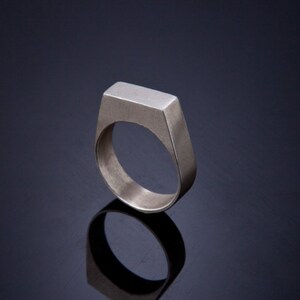 May include: A silver ring with a flat, rectangular top. The ring is simple and modern in design.