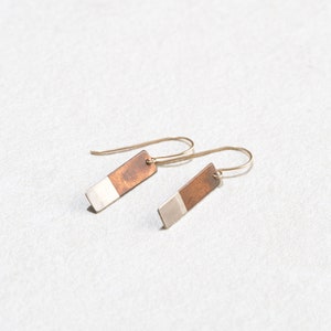 以下が含まれることがあります： 長方形の銀と銅のイヤリングのペア。イヤリングは細い金線から吊り下げられています。
