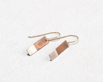 Op de afbeelding: Een paar rechthoekige oorbellen van zilver en koper. De oorbellen hangen aan een dunne gouden draad.