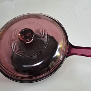 May include: A round, purple-tinted glass skillet with a matching glass lid. The lid features a central knob and a radiating sunburst pattern etched into the glass. The skillet has a long, integrated glass handle extending from one side.