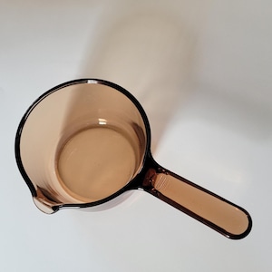 May include: A brown glass measuring cup with a handle and a pouring spout. The cup is empty and sits on a white surface. The handle is rectangular and the cup is round.