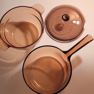 May include: A set of vintage amber-colored glass cookware, including a round casserole dish with handles, a lid with a knob, and a skillet with a handle. The pieces are arranged on a light surface, showcasing their transparent design.