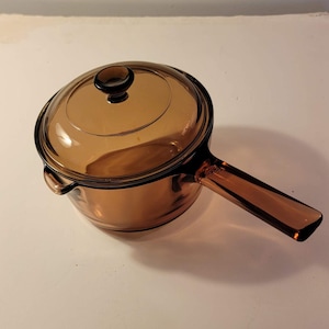 May include: A vintage amber-colored glass saucepan with a matching lid. The saucepan has a long, rectangular handle and a rounded body. The lid has a small, round knob. This cookware is ideal for cooking and serving.