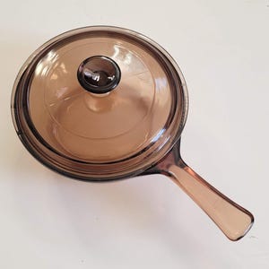 May include: A brown glass saucepan with a matching lid. The pan has a handle and is sitting on a white surface. The lid has a dark knob.