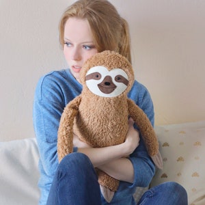 May include: A young woman wearing blue jeans and brown work boots is holding a brown plush sloth with a white face and brown eyes.