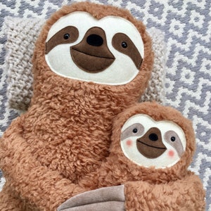 May include: Two plush brown sloth toys, one larger than the other, are hugging. The larger sloth has a white face with brown eyes and a brown nose. The smaller sloth has a white face with brown eyes and a brown nose. Both sloths have a brown mouth.