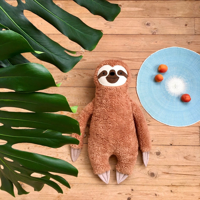 May include: A brown plush sloth toy with a white face and black eyes. The sloth is lying on a wooden surface with a blue and white patterned circle behind it.