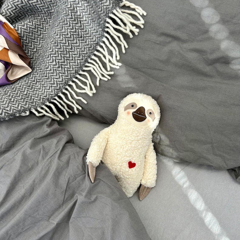 May include: A white plush sloth toy with a brown nose and a red heart on its chest. The sloth is lying on a gray blanket.