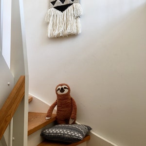 May include: A brown sloth plush toy sitting on a black and white patterned cushion on wooden stairs. A black and white geometric wall hanging with white fringe is above the stairs.