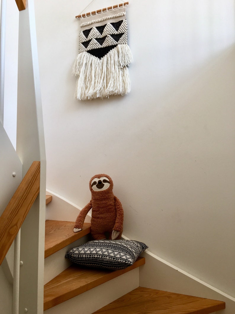 May include: A brown sloth plush toy sitting on a black and white patterned cushion on wooden stairs. A black and white geometric wall hanging with white fringe is above the stairs.