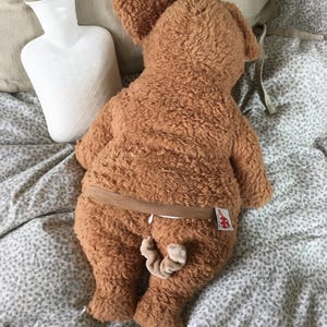 May include: A brown plush dog-shaped hot water bottle with a tail and a brown elastic band around the middle. A white hot water bottle is next to the plush dog on a bed with a patterned comforter.