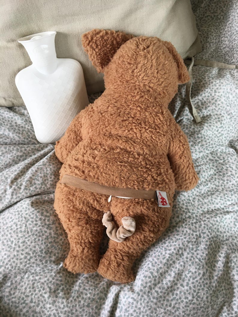May include: A brown plush dog-shaped hot water bottle with a tail and a brown elastic band around the middle. A white hot water bottle is next to the plush dog on a bed with a patterned comforter.