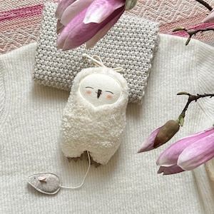 Pode incluir: Um brinquedo de coruja de pelúcia branca com um rosto sorridente e uma corda presa a um pequeno brinquedo de rato cinza. A coruja está deitada em um suéter branco, com um cachecol de malha cinza e flores de magnólia rosa no fundo.