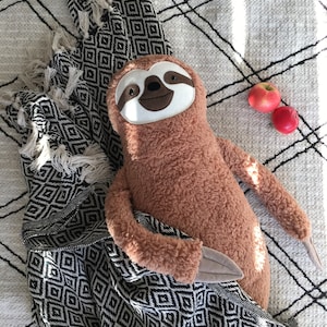 May include: A brown plush sloth toy with a white face and black eyes, lying on a black and white patterned blanket with red apples in the background.