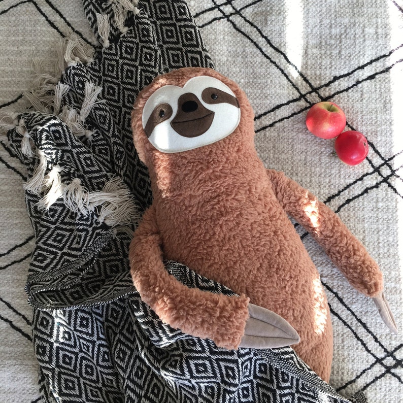 May include: A brown plush sloth toy with a white face and black eyes, lying on a black and white patterned blanket with red apples in the background.