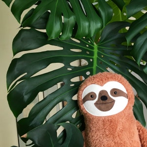 May include: A plush brown sloth toy with a white face and brown eyes. The sloth is sitting in front of a green monstera plant.