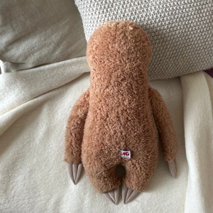 May include: A brown plush sloth toy with a small red tag on its back. The sloth is lying on a white blanket.
