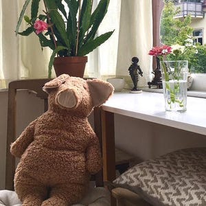 May include: A plush, brown teddy bear with large ears sits on a chair. A potted plant with green leaves and pink flowers is in the background. A glass of water with flowers and a small statue are on a white table.