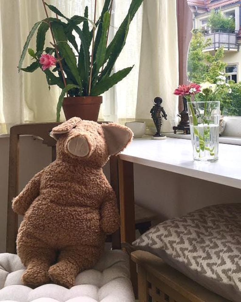May include: A plush, brown teddy bear with large ears sits on a chair. A potted plant with green leaves and pink flowers is in the background. A glass of water with flowers and a small statue are on a white table.