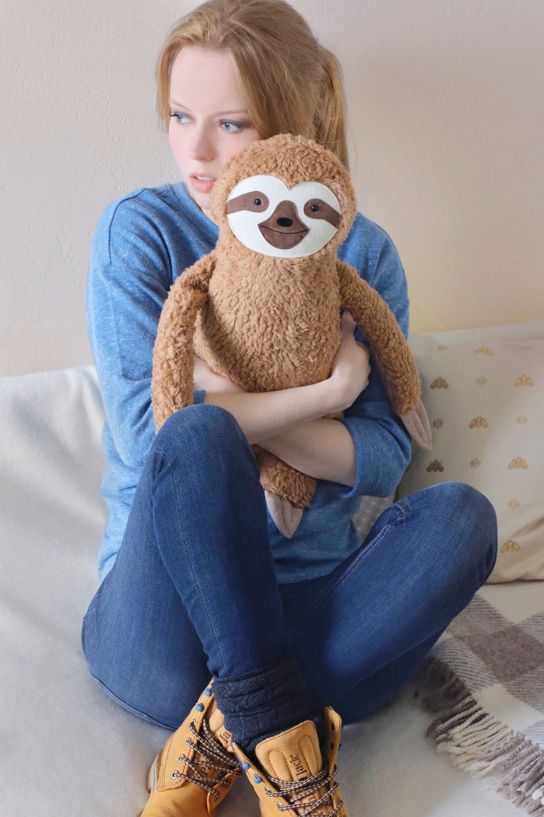 May include: A young woman wearing blue jeans and brown work boots is holding a brown plush sloth with a white face and brown eyes.