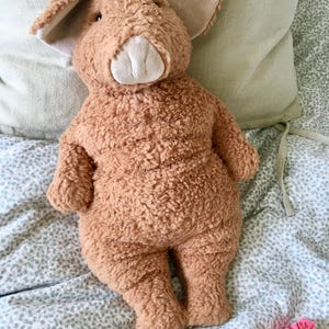 May include: A plush, brown pig stuffed animal with large ears and a white snout. The pig is made of a soft, textured material and is lying on a bed with a patterned comforter and a light green pillow.