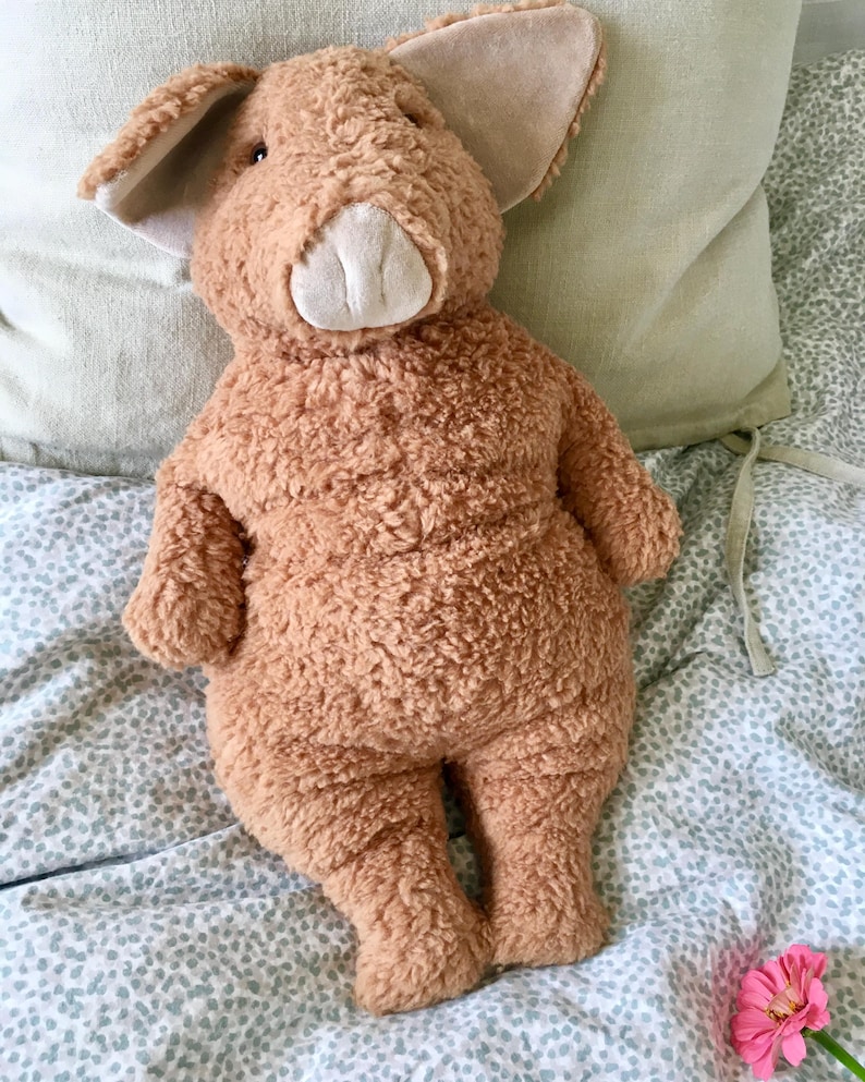 May include: A plush, brown pig stuffed animal with large ears and a white snout. The pig is made of a soft, textured material and is lying on a bed with a patterned comforter and a light green pillow.