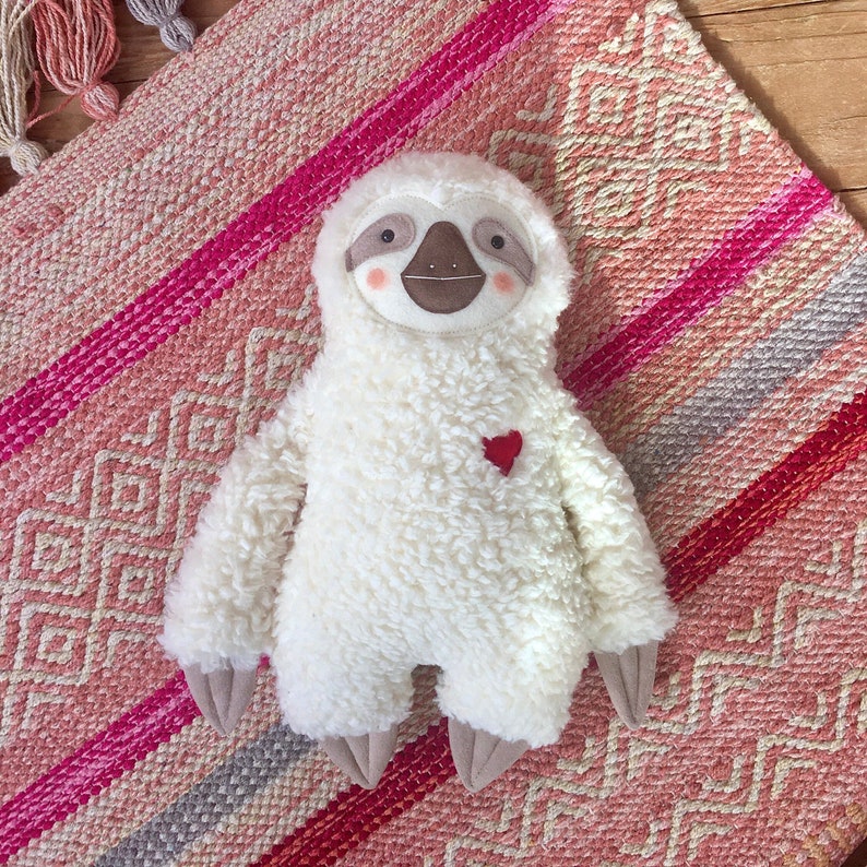 May include: A white plush sloth toy with a brown face and a red heart on its chest. The sloth is lying on a pink and white patterned rug.