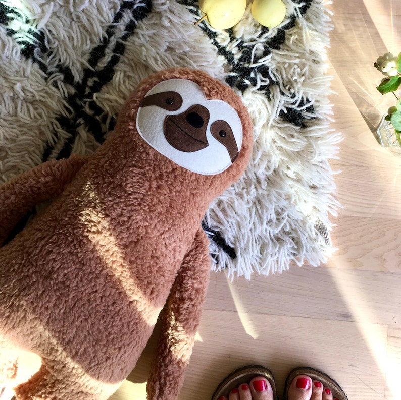 May include: A brown plush sloth toy with a white face and brown eyes. The toy is lying on a white and black fluffy rug.