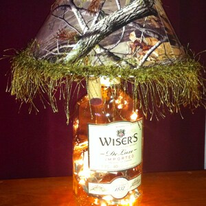 May include: A unique lamp made from a whiskey bottle, featuring a camouflage lampshade with a decorative fringe. The bottle is filled with fairy lights, and the label reads "Wiser's De Luxe".