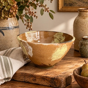 May include: A hand-thrown ceramic bowl with a warm tan glaze, featuring irregular cream and pale yellow painted accents on the exterior and interior. The bowl has a wide, open rim and a tapered base, showcasing a smooth, glossy finish.