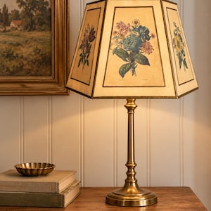 May include: A table lamp with a brass-toned metal base and a hexagonal lampshade. The shade features cream-colored panels, each decorated with a distinct botanical illustration of colorful flowers, framed by thin dark brown border lines.