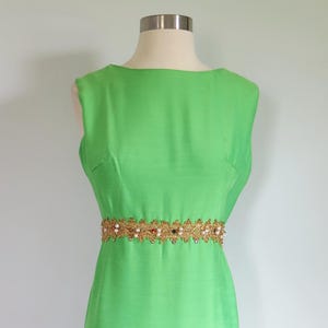 Puede incluir: Vestido sin mangas verde lima con cinturón dorado y adornado con perlas. El vestido se exhibe en un maniquí. El vestido es de largo midi y la tela tiene un brillo sutil. El cinturón es un adorno decorativo.