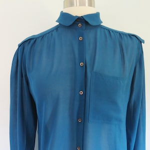 May include: A teal blue blouse with a rounded collar and long sleeves. The blouse features a front pocket and button-down closure with small, round buttons. The shoulders have a slight padded detail. The fabric appears to be semi-sheer.