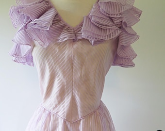 Può includere: Un abito color lavanda chiaro con scollo a V e maniche corte con volant pronunciati. Il tessuto presenta una sottile trama a righe diagonali su tutto il corpetto e la gonna, con una cucitura in vita a punta che definisce la struttura.