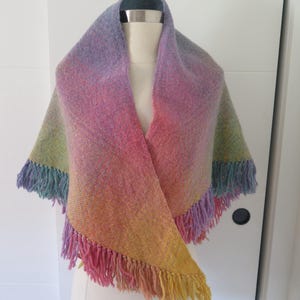 May include: A triangular woven shawl featuring a gradient of colors including yellow, orange, pink, purple, blue, and green. The fabric has a textured weave and is finished with long, matching fringe along the bottom edges.