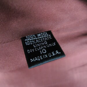 Pu&ograve; includere: Un'etichetta in tessuto nero con testo bianco che recita "100% Lana 100% Acetato Fodera Lavaggio a secco solo 10 Made in USA."