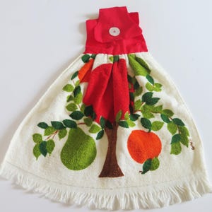 May include: A hanging kitchen towel with a red fabric top featuring a white button closure. The cream-colored towel has a fringed bottom edge and a printed design of a brown tree with green leaves, a green pear, and an orange fruit.