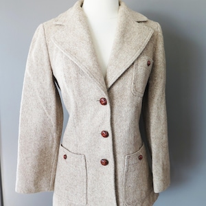 Pode incluir: Blazer de tweed bege com lapelas entalhadas, dois bolsos frontais e botões marrons. O blazer é exibido em um manequim e tem um design clássico e sob medida. O tecido tem uma aparência texturizada.