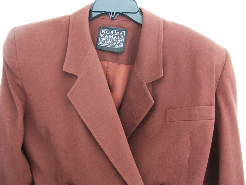 Pu&ograve; includere: Un blazer marrone con chiusura a un solo bottone e una tasca sul lato destro. L'etichetta recita "Norma Kamali The Exclusive Collection for Bloomingdales".