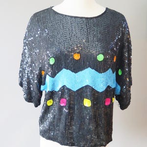 Puede incluir: Top negro con lentejuelas, cuello redondo y mangas cortas. El top presenta un diseño en zigzag azul y lunares de colores naranja, verde, rosa y amarillo. La prenda se exhibe en un maniquí.