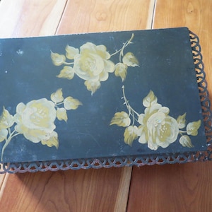 May include: A rectangular black metal box featuring a hand-painted floral design of three pale yellow roses with green leaves. The box is surrounded by a decorative, scalloped metal filigree border that extends around all four sides of the lid.