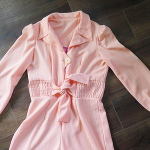 Könnte beinhalten: Hellpfirsichfarbener Jumpsuit mit langen Ärmeln und Kragen. Die Taille ist mit einem gerafften Detail und einem passenden Bindegürtel versehen. Ein einzelner Knopf ist sichtbar. Der Jumpsuit ist aus weichem, fließendem Stoff.