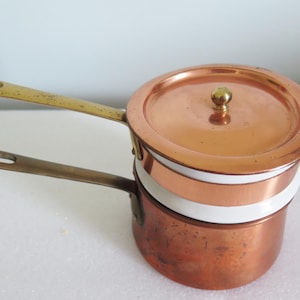 May include: Two copper saucepans with brass handles and a copper lid. The pans are stacked, with a white band separating them. The lid has a small brass knob. The copper has a warm, reddish-brown hue, suggesting vintage cookware.